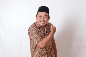 Portrait of excited Asian man wearing batik shirt and songkok smiling and looking at camera, making thumbs up hand gesture. Isolated image on gray background photo