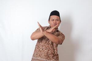 Portrait of excited Asian man wearing batik shirt and songkok  showing X sign of hands, refusing an invitation from someone, making prohibitive gesture. Isolated image on gray background photo