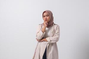 Portrait of confused Asian hijab woman in casual suit thinking with hand on chin, feeling doubt while making choice. Businesswoman concept. Isolated image on white background photo