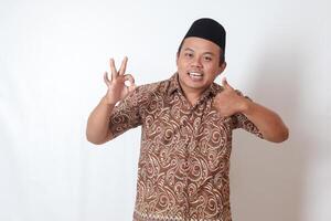 Portrait of excited Asian man wearing batik shirt and songkok showing ok hand gesture and smiling looking at camera. Isolated image on gray background photo