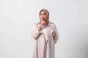 Portrait of excited Asian hijab woman in casual suit making hush gesture and asking to be quiet. Businesswoman concept. Isolated image on white background photo