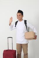 Portrait of happy Asian muslim man carrying cardboard box and suitcase while saying hi to greet someone. Going home for Eid Mubarak. Isolated image on white background photo