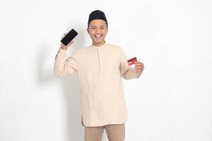 Portrait of attractive Asian muslim man in koko shirt with skullcap holding a credit card while showing empty screen on mobile phone for mock up and graphic. Isolated image on white background photo