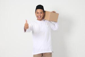 Portrait of excited Asian muslim man in koko shirt with peci carrying cardboard box while pointing and showing thumb up hand gesture. Going home for Eid Mubarak. Isolated image on white background photo