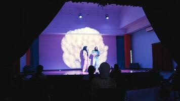 Two women in hijabs performing music, one singing into a microphone and the other playing a ukulele, with stage lighting and shadows in the background. video