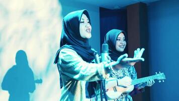 Two women in hijabs performing music, one singing into a microphone and the other playing a ukulele, with stage lighting and shadows in the background. video