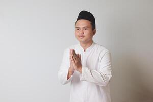 Portrait of Asian muslim man in white koko shirt showing apologize and welcome hand gesture. Isolated image on gray background photo