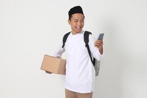 Portrait of happy Asian muslim man holding mobile phone while carrying cardboard box with excited expression. Going home for Eid Mubarak. Isolated image on white background photo