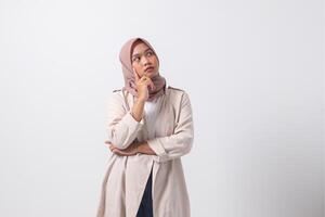 Portrait of confused Asian hijab woman in casual suit thinking with hand on chin, feeling doubt while making choice. Businesswoman concept. Isolated image on white background photo