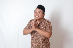 Portrait of smiling Asian man wearing batik shirt and songkok showing apologize and welcome hand gesture. Ramadan advertising concept. Isolated image on gray background photo