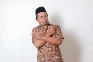 Portrait of confused Asian man wearing batik shirt and songkok with crossed hands, pointing sideways, making choice, choosing between two objects. Isolated image on gray background photo