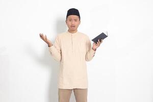 Portrait of confused Asian muslim man in koko shirt with peci difficulty understanding the contents of the book, reading a textbook. Isolated image on white background photo