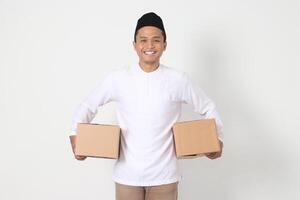 Portrait of attractive Asian muslim man in koko shirt with peci feeling grateful and peacful while carrying cardboard box. Going home for Eid Mubarak. Isolated image on white background photo