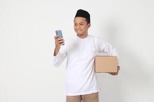 Portrait of excited Asian muslim man in koko shirt with peci carrying cardboard box while holding mobile phone. Going home for Eid Mubarak. Isolated image on white background photo