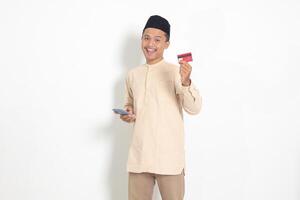 Portrait of attractive Asian muslim man in koko shirt with skullcap holding a mobile phone and presenting credit card. Isolated image on white background photo