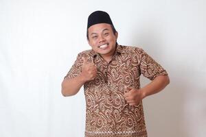 Portrait of excited Asian man wearing batik shirt and songkok smiling and looking at camera, making thumbs up hand gesture. Isolated image on gray background photo