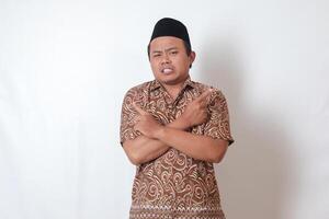 Portrait of confused Asian man wearing batik shirt and songkok with crossed hands, pointing sideways, making choice, choosing between two objects. Isolated image on gray background photo