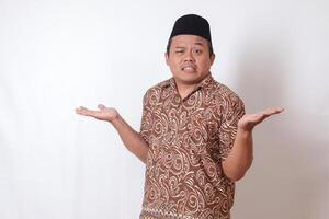 Portrait of confused Asian man wearing batik shirt and songkok with crossed hands, pointing sideways, making choice, choosing between two objects. Isolated image on gray background photo