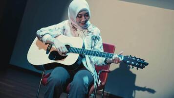 sorridente mulher dentro hijab jogando acústico guitarra enquanto sentado em uma cadeira, com uma caloroso e acolhedor ambiente. video