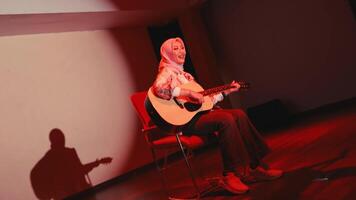 sonriente mujer en hijab jugando acústico guitarra mientras sentado en un silla, con un calentar y acogedor ambiente video