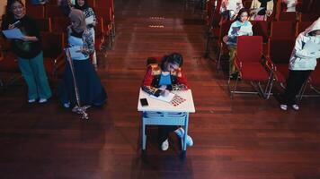 niño sentado a un escritorio en un sala con un atento audiencia en el antecedentes. video