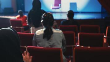 Publikum Mitglieder sitzend im ein schwach zündete Auditorium gegenüber ein Bühne, Erfassen das Konzept von ein Leben Performance oder Fall. video