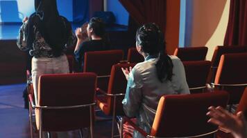 Audience members seated in a theater with focus on empty red chairs. video