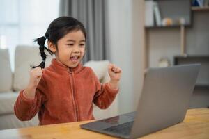 Young Girl Celebrating Success on Laptop at Home. Joyful young Asian girl with raised fists in celebration while using a laptop at a wooden table. Education for kid student concept. photo