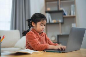 Concentrated Asian young girl studying on laptop at home. Young Asian girl focused on using a laptop for her studies in a comfortable home setting. Education for kid student concept. photo