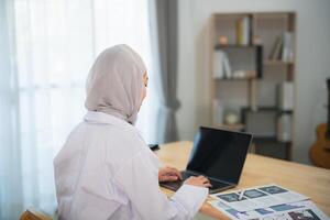 Back view. Muslim islam Asian doctor man wearing hijab and stethoscope working with laptop computer, smartphone. Female doctor work at home office hospital. Health hospital concept. photo