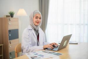 Muslim islam Asian doctor man wearing hijab and stethoscope working with laptop computer, smartphone. Female doctor work at home office hospital. Health hospital concept. photo