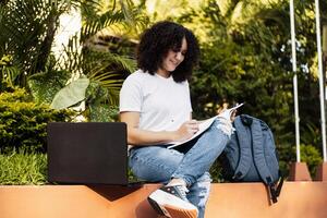 joven estudiante con Rizado pelo haciendo notas en un libro, latín americano hembra estudiante haciendo tarea. foto