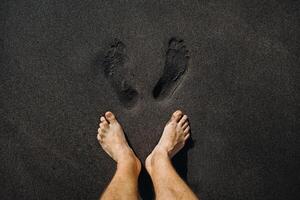 Close up of male footprints and feet walking on the volcanic black sand on the beach photo