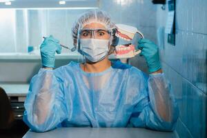 un dental médico vistiendo azul guantes y un máscara sostiene un dental modelo de el Superior y inferior mandíbulas y un dental espejo foto