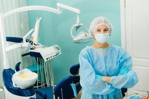 un hembra dentista vistiendo un médico máscara y caucho guantes poses para el cámara y pliegues su brazos en su oficina foto