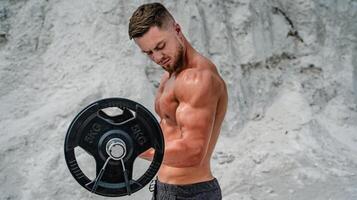 Brutal strong bodybuilder posing outdoor. Photoshoot in a quarry. Outdoor sports concept. Training with dumbbell. Posing and pumping muscles. White landscape. photo