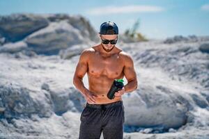 retrato de un hermoso medio desnudo carrocero hombre en lentes y un gorra con un botella de agua. foto