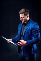 Half-length portrait of handsome young smiling man wearing dark shirt and blue suit holding blue folder, looking at camera. Black background. Money and business concept. photo