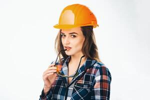 Happy young woman is wearing orange safety helmet and checkered shirt. Looking at camera, yellow safety glasses near lips. Isoalted background. photo
