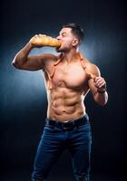 Athlete with naked torso eats fresh bread. Sausages on a neck. Cropped photo. Lifestyle concept. Studio photo. Hungry sportsman. photo