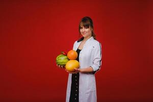 un hembra médico enfermero en un blanco Saco con Fruta en su manos poses en un rojo fondo, melón, sandía, fresa y pomelo foto