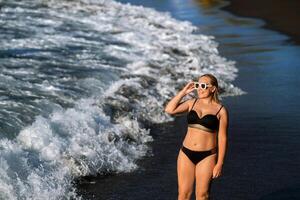 un niña en un negro traje de baño camina en el playa en el isla de tenerife en el atlántico océano, España foto