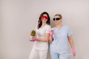 Two cosmetologists in protective glasses are holding a laser depilation device and a cactus in their hands photo