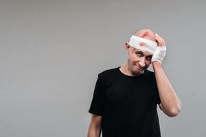 A battered and battered man in a black T-shirt stands against a gray background, holding his aching head with his hands wrapped around it photo