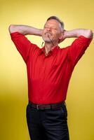 hermoso medio años mayor hombre vistiendo un rojo camisa terminado aislado amarillo antecedentes. relajante y extensión con brazos y manos detrás cabeza y cuello. foto