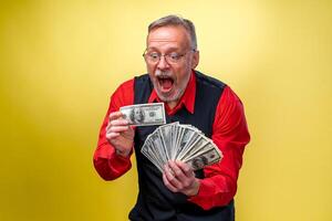 Portrait of happy and white teeth smile senior old businessman. Man holding money in hands. Dressed in red shirt, isolated on yellow background. Human emotions and facial expressions photo