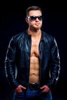 Young handsome man in sunglasses with open leather jacket revealing muscular chest and torso. Male looking side. Closeup photo