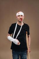 A battered man with a bandaged head and a cast on his arm stands on crutches on a gray background photo