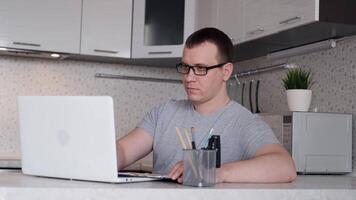 joven masculino ingeniero en lentes trabajos desde hogar sentado a un mesa utilizando un ordenador portátil video