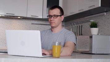 Jeune Masculin pigiste dans des lunettes les usages une portable tandis que travail de Accueil sur le l'Internet, à la recherche à le filtrer, souriant tandis que travail à distance tandis que séance à une table video
